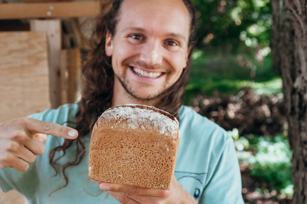 Jannis von AHO im grünen Garten, hält ein Sprossen Brot in der Hand und lacht in die Kamera